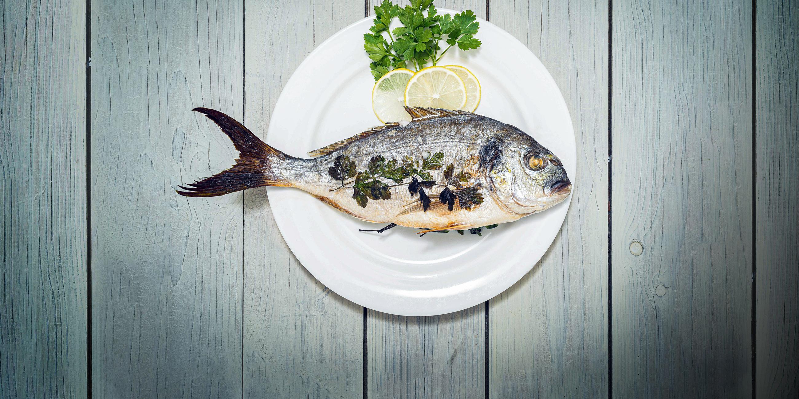 Nahaufnahme von Fisch (Seebrasse) mit Petersilie und Zitrone in Teller auf Holztisch.