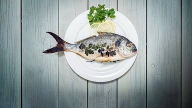 Nahaufnahme von Fisch (Seebrasse) mit Petersilie und Zitrone in Teller auf Holztisch.