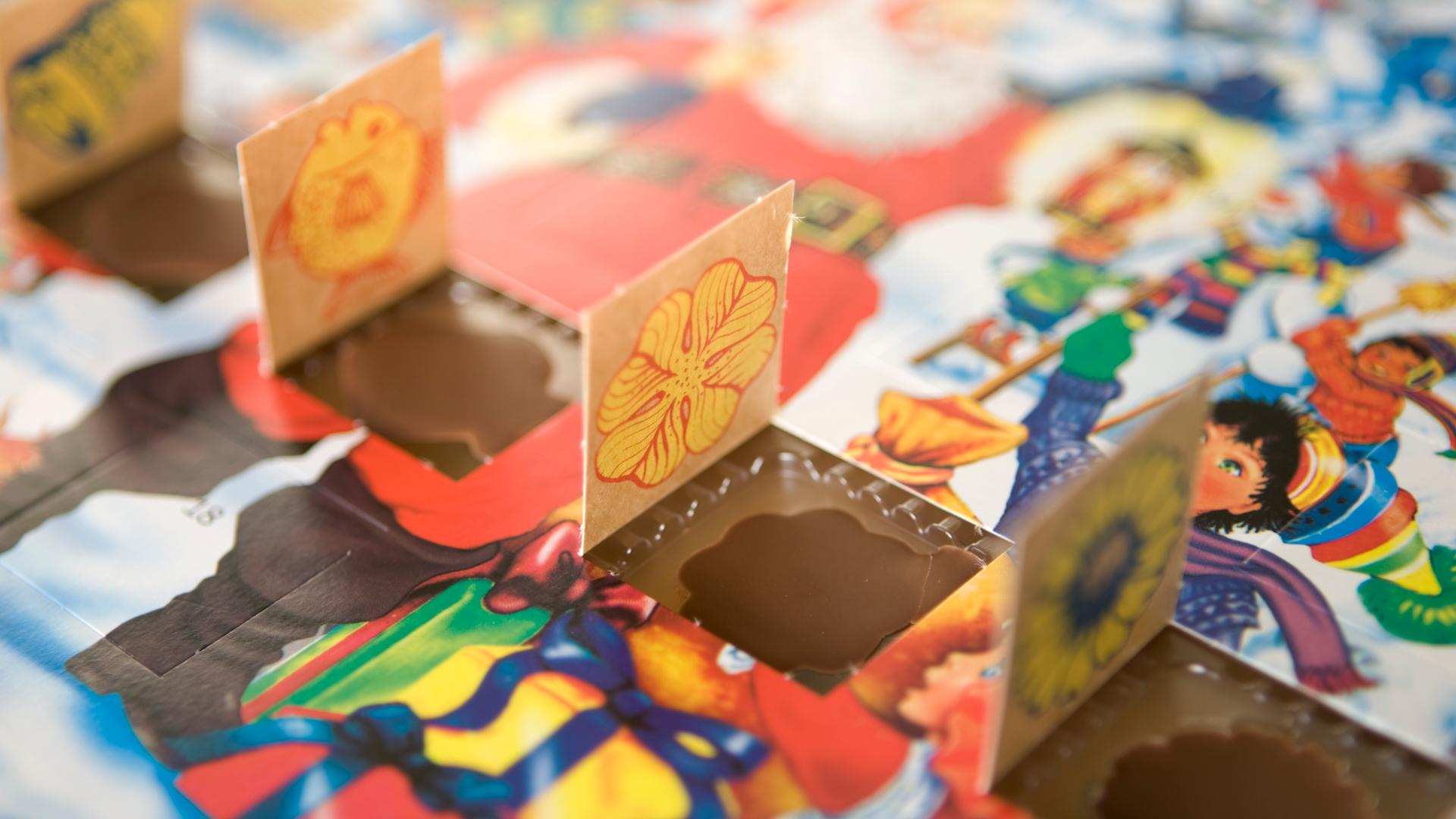 Man sieht einen Adventskalender mit winterlichem Motiv an dem vier Türchen nebeneinander geöffnet sind. Die kleinen Schokoladenmotive liegen noch in ihren Förmchen. 