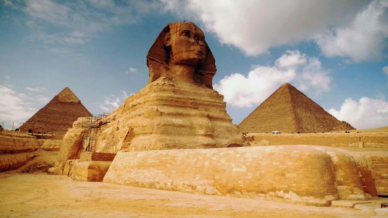 Schrägansicht von unten auf die große Sphinx von Gizeh. An der linken Seite der Sphinx steht ein Baugerüst. Der Himmel ist bewölkt. Im Hintergrund sind zwei Pyramiden zu erkennen.