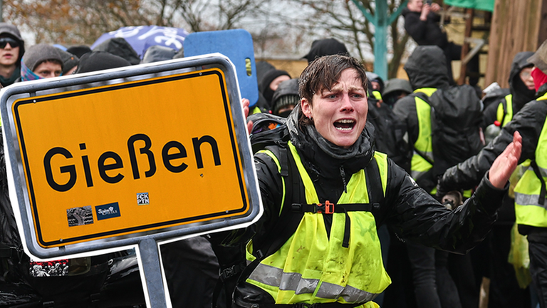 Eine Frau mit Warnweste in einer Menge Protestierender und ein Ortsschild von Gießen. 