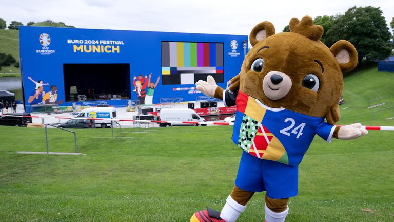 Albärt, das Maskottchen der Fußball-EM 2024, steht im Olympiapark vor einer Leinwand und einer Bühne für die Fan Zone