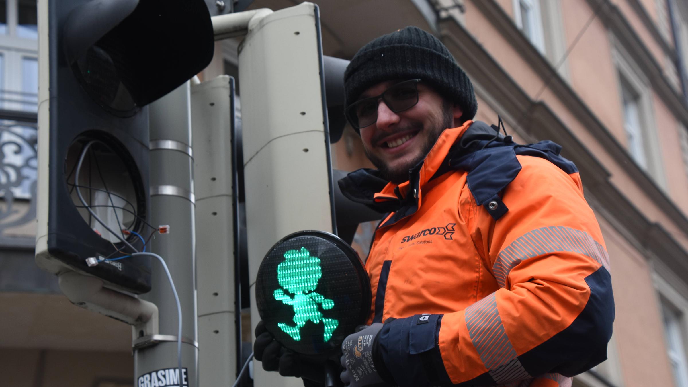 Ein Mann in orangener Warnjacke baut eine Ampel zusammen. Das Ampelmännchen ist eine Pumuckl-Figur.
