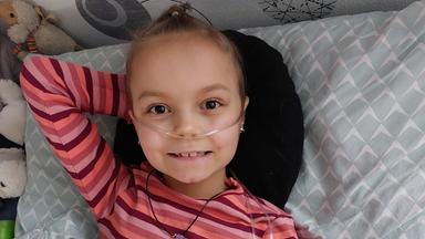 Mädchen liegt im Bett und lächelt, in ihrer Nase steckt ein Schlauch. 