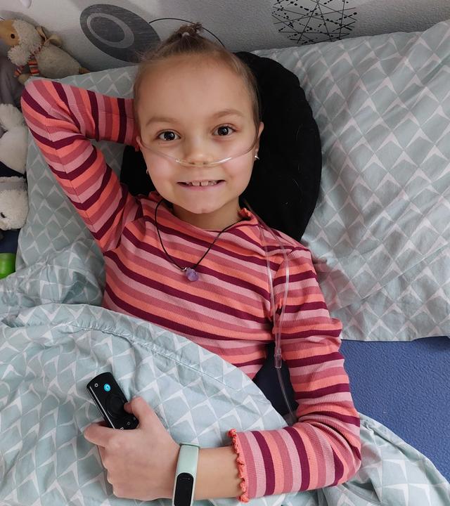 Mädchen liegt im Bett und lächelt, in ihrer Nase steckt ein Schlauch. 