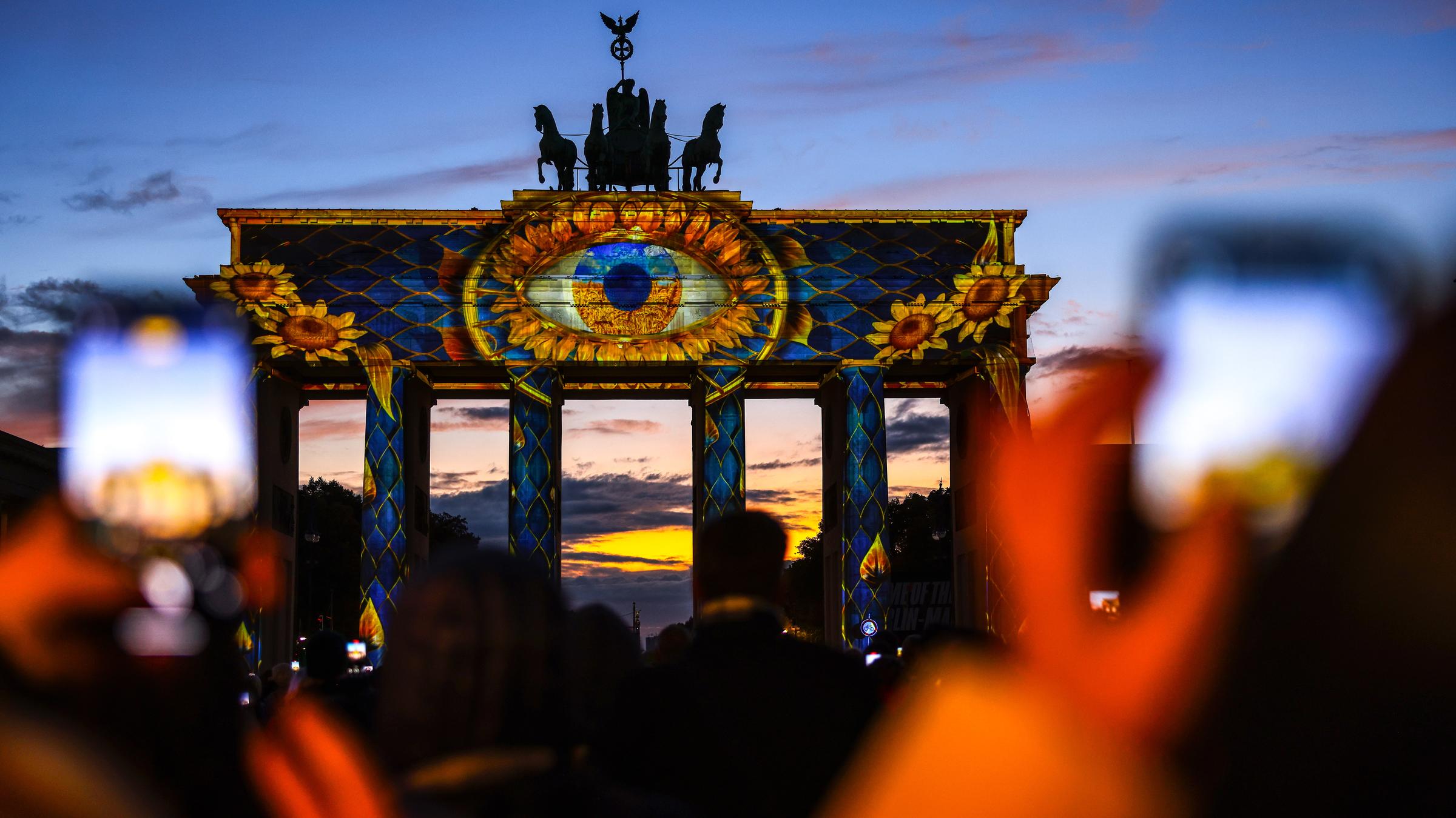 Das Brandenburger Tor wird angeleuchtet, zu sehen ist ein großes Auge. Hinten geht die Sonne unter.