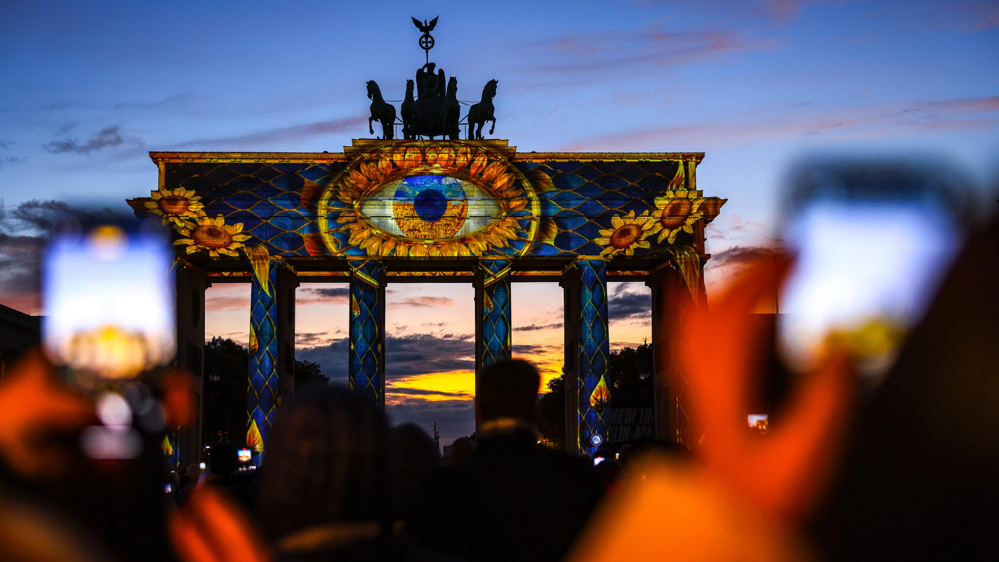 Das Brandenburger Tor wird angeleuchtet, zu sehen ist ein großes Auge. Hinten geht die Sonne unter.