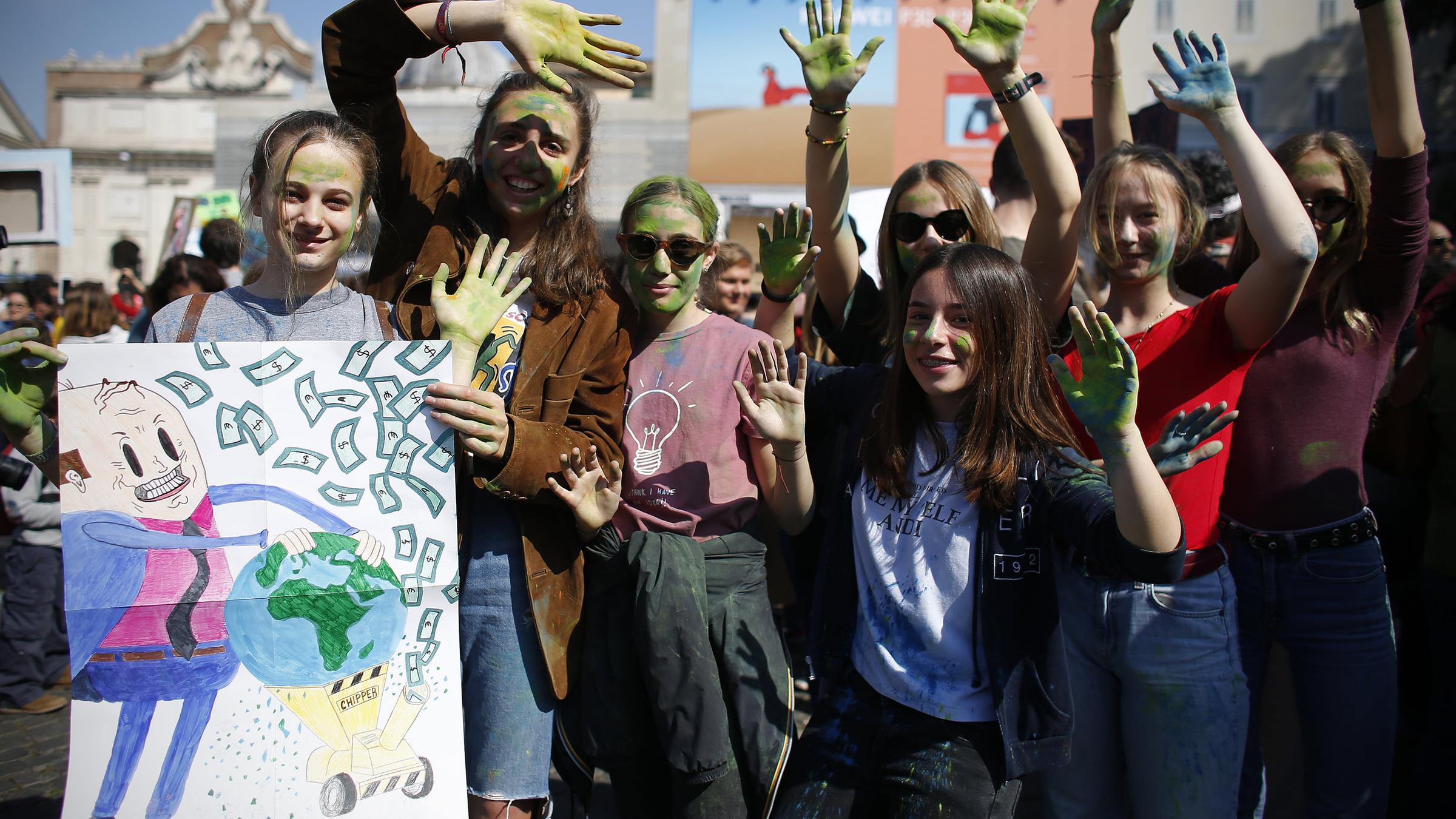 Junge Demonstrantinnen halten blau-grün bemalte Hände in die Luft und halten ein Plakat mit einer Zeichnung hoch, auf dem ein Mann im Anzug die Erde in einen Hächsler steckt und aus ihr Geld macht.