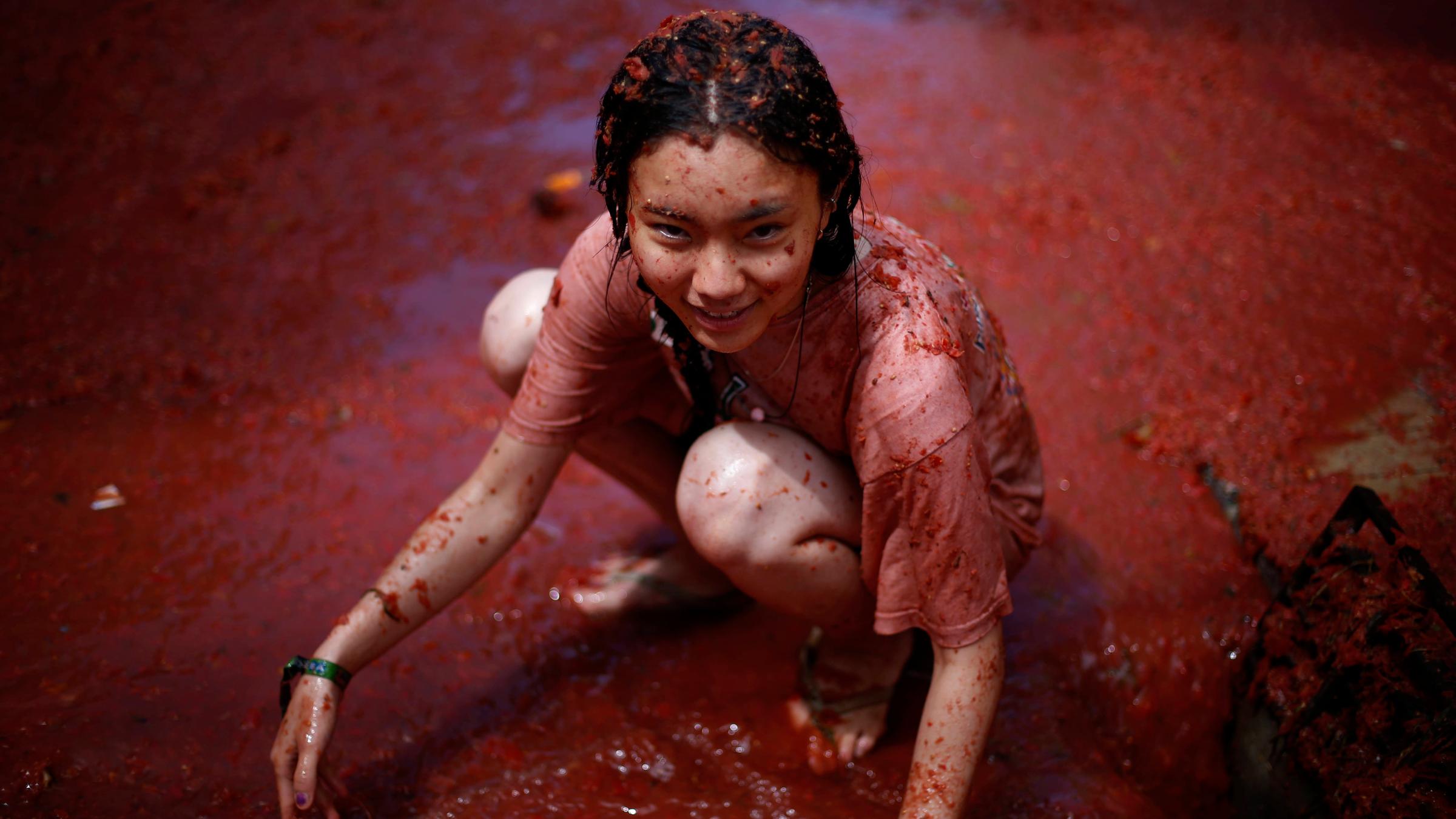 Ein Mädchen kniet in einer riesigen Pfütze roter Matsche. sie ist von Kopf bis Fuß mit Tomaten begossen, denn sie ist Teil der Tomatenschlacht.