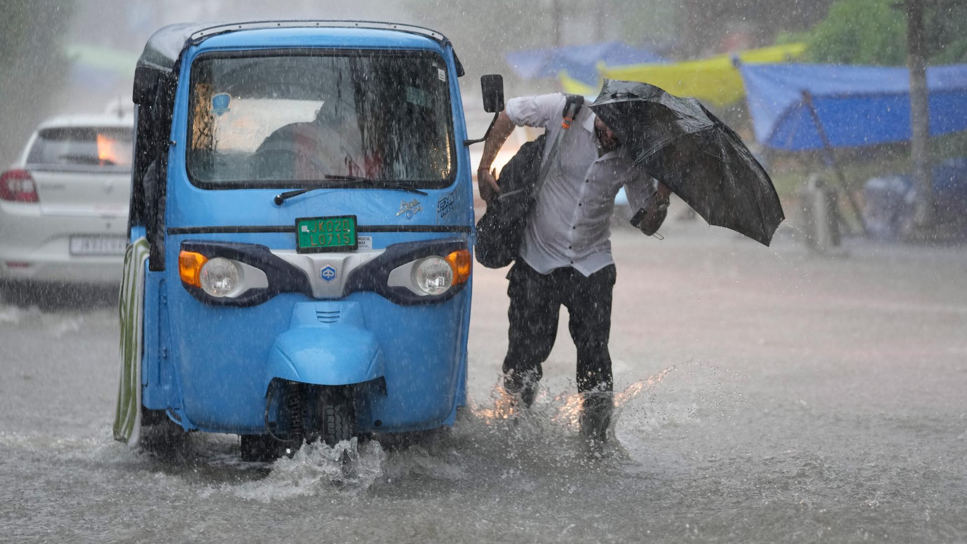 Blauer Waagen auf drei Rädern - ein TukTuk und ein Mensch mit Regenschirm im Monsunregen.