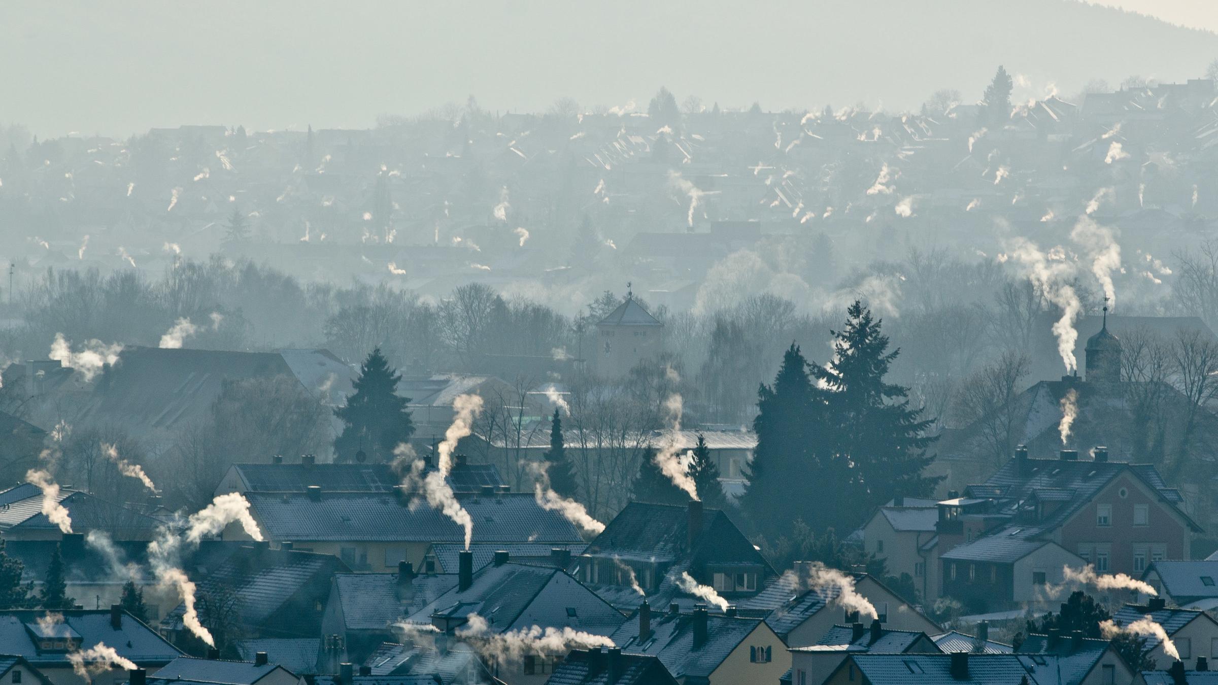 In Bayern rauchen die Schornsteine in einem Wohngebiet. Die Dächer der Häuser sind mit Schnee bedeckt.