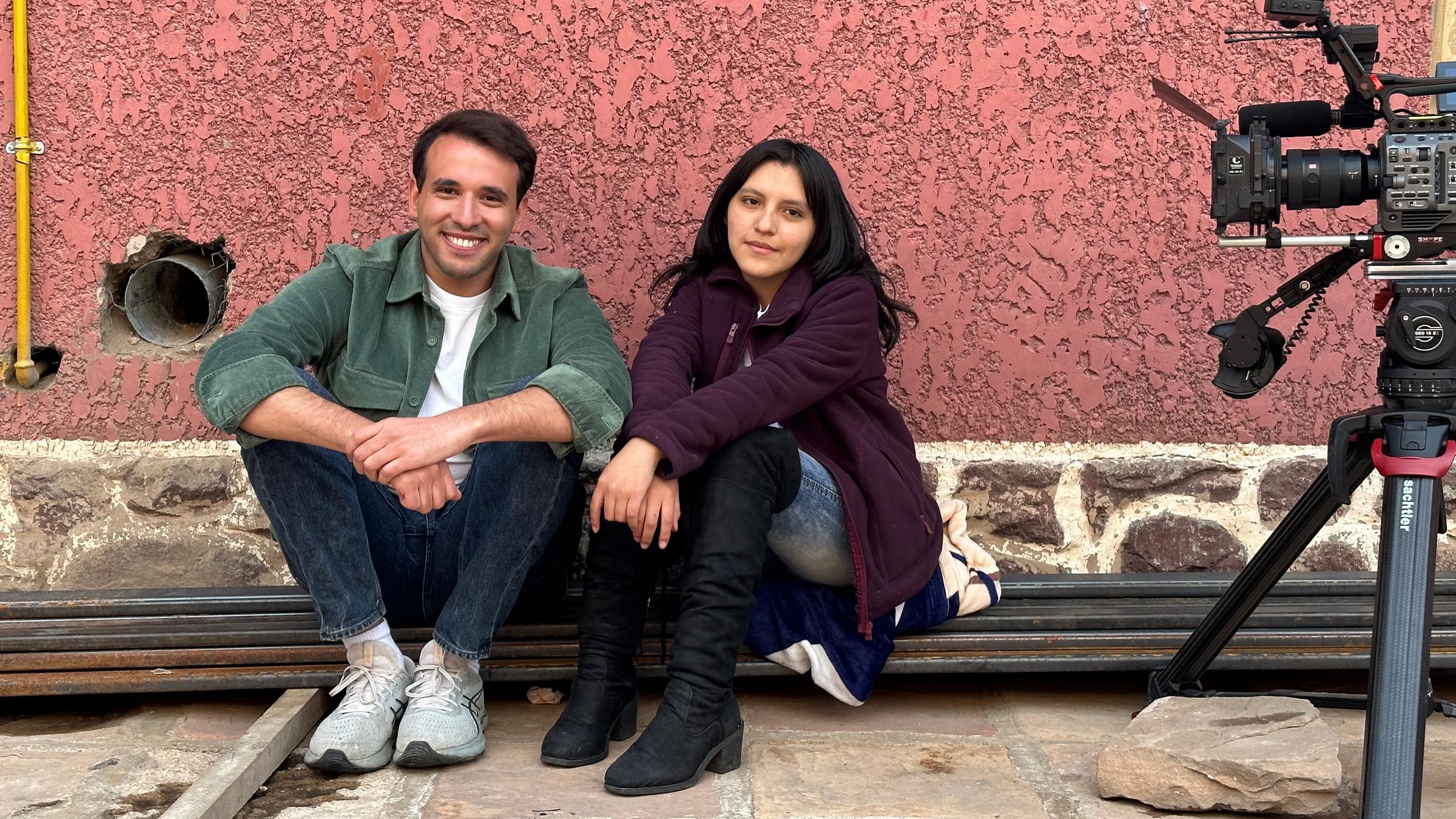 logo!-Reporter Sherif sitzt naben Mädchen auf Schienen vor roséroter Fassade