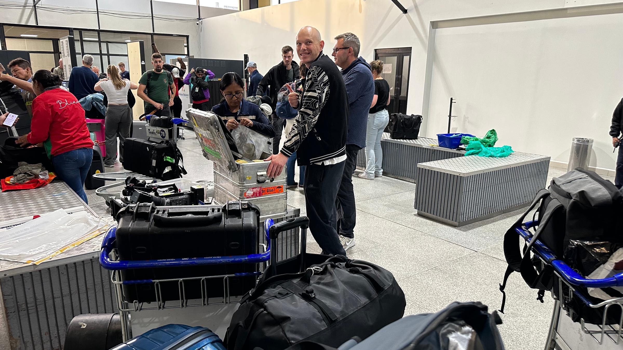 Eine Metallkiste wird von Frau bei Gepäckkontrolle im Flughafen geöffnet. Im Vordergrund Gepäck, im Hintergrund stehen Menschen mit Gepräck Schlange