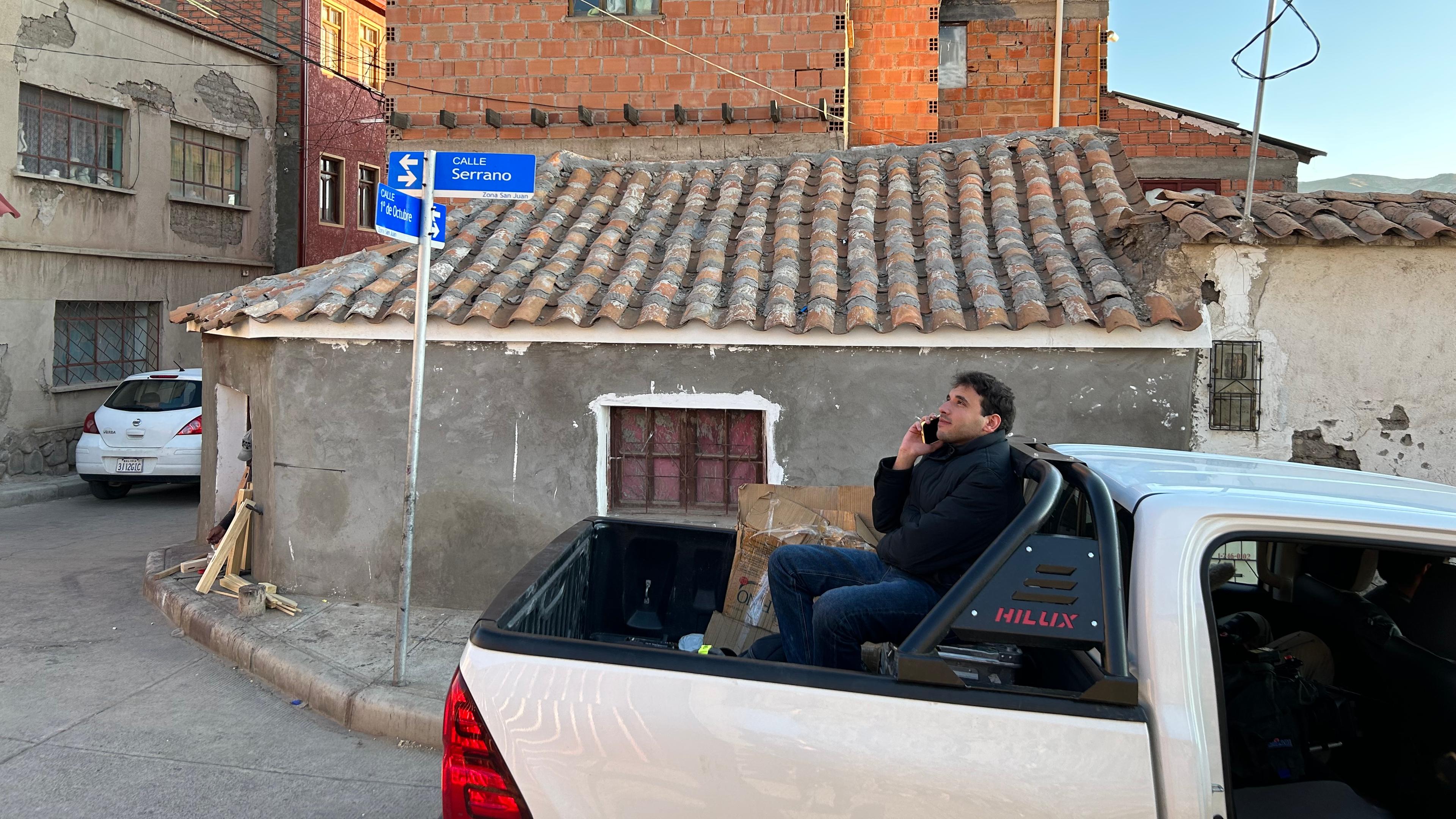 Ladefläche von Pick-up, auf der logo!-Reporter Sherif sitzt, als Seitenaufnahme. Mit Straßenschild und Häusern im Hintergrund.