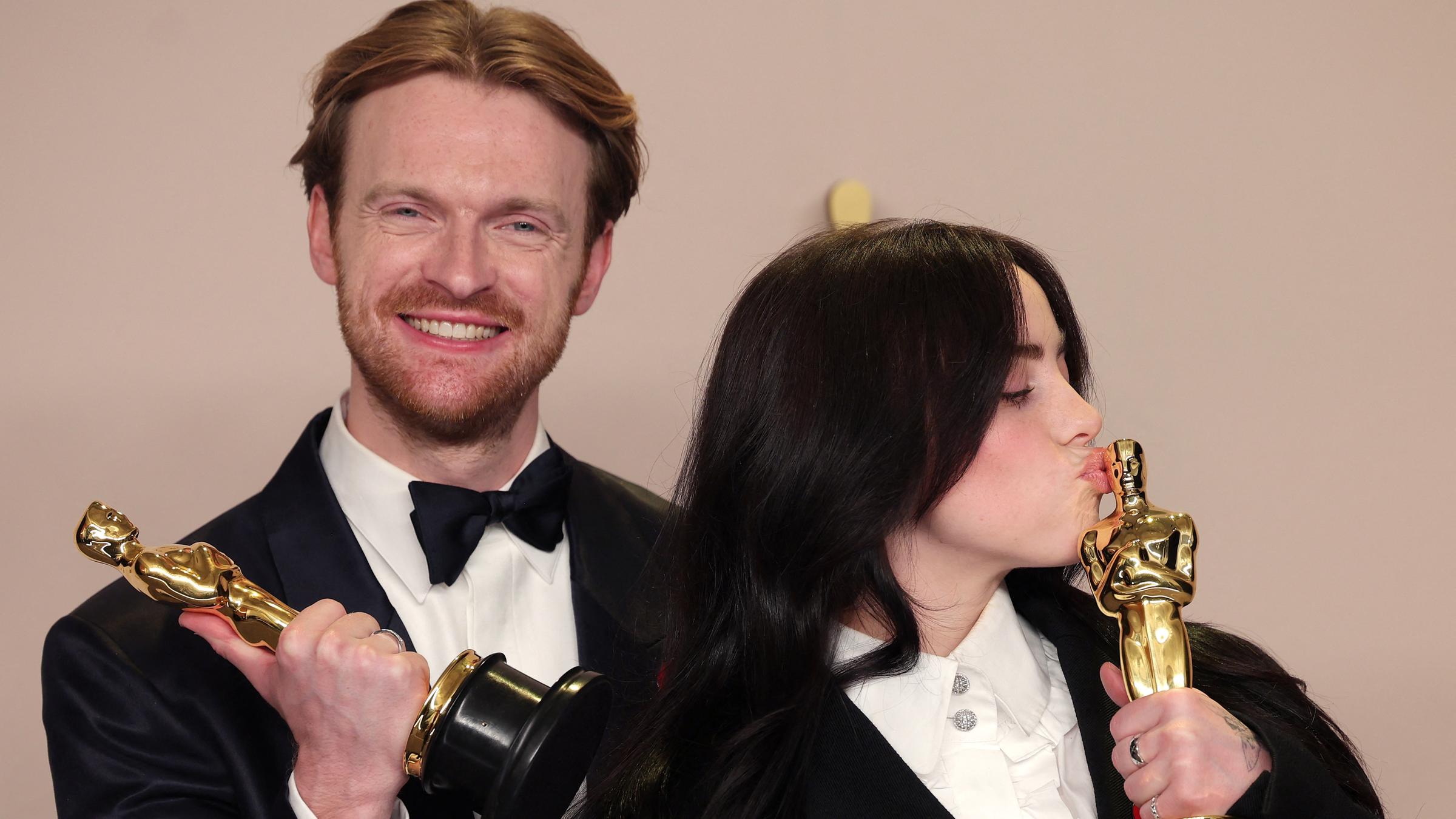 Billie Eilish küsst ihren Oscar. Neben ihr steht Finneas O'Connell, ihr Bruder und hält lächeln einen Oscar in der Hand.