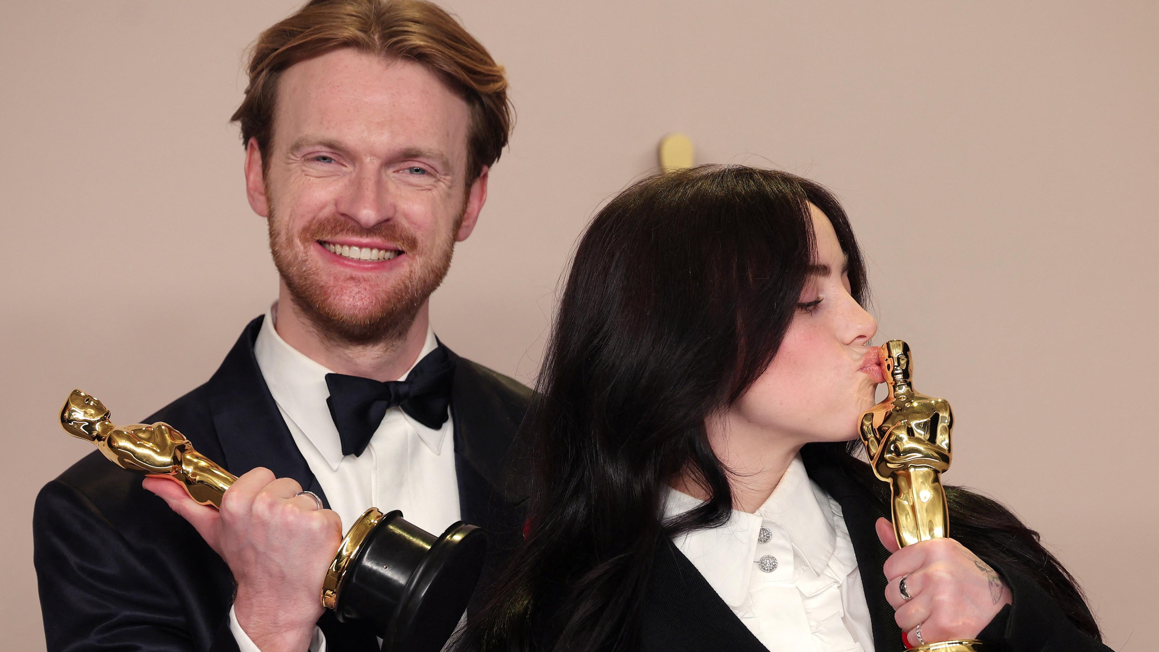 Billie Eilish küsst ihren Oscar. Neben ihr steht Finneas O'Connell, ihr Bruder und hält lächeln einen Oscar in der Hand.