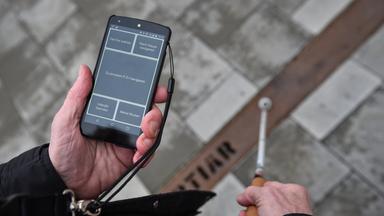 Mann hält Handy mit Verkehrsapp für Blinde und Blindenstock in Hand
