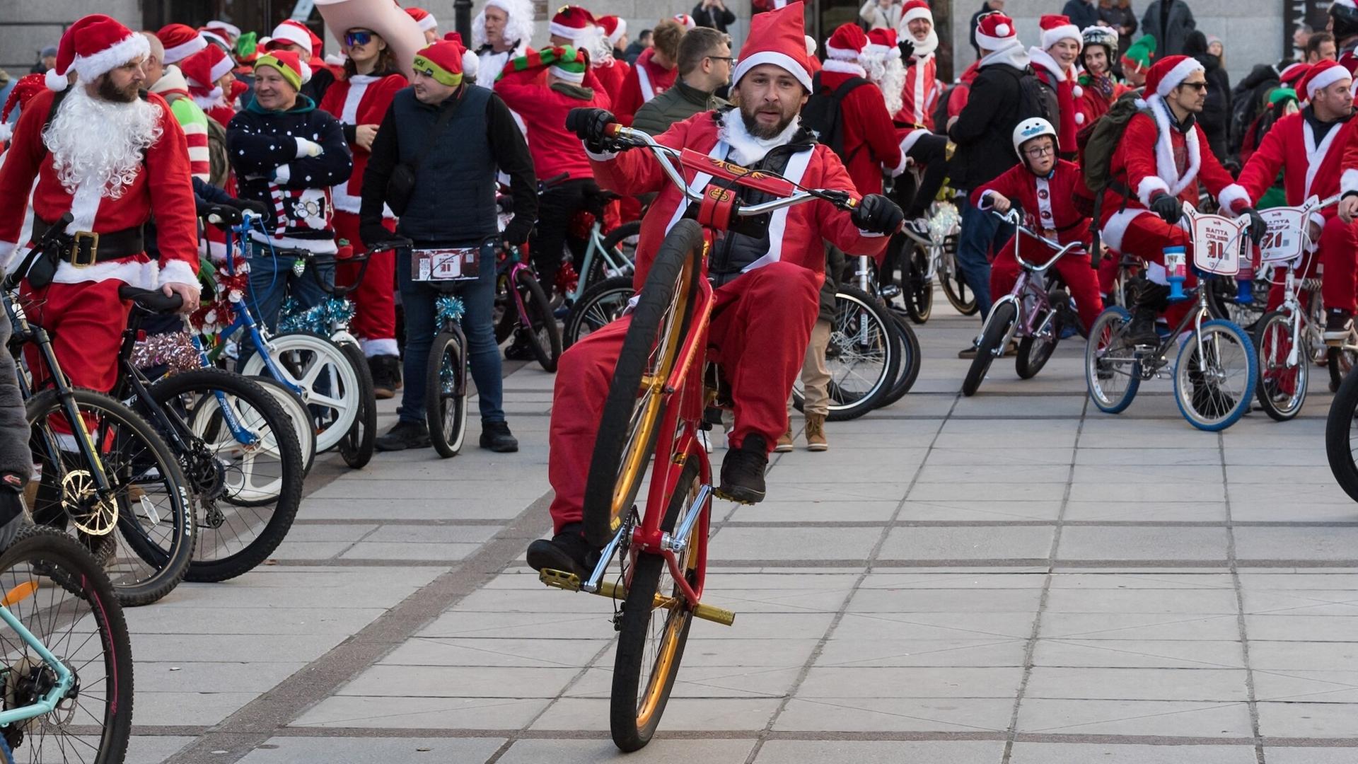 Im Vordergrund fährt ein Mann im Weihnachtsmannkostüm auf einem BMX-Rad und hebt dabei sogar das Vorderrad an. Dahinter rollen viele weitere verkleidete Weihnachtsmänner gemeinsam durch die Stadt.