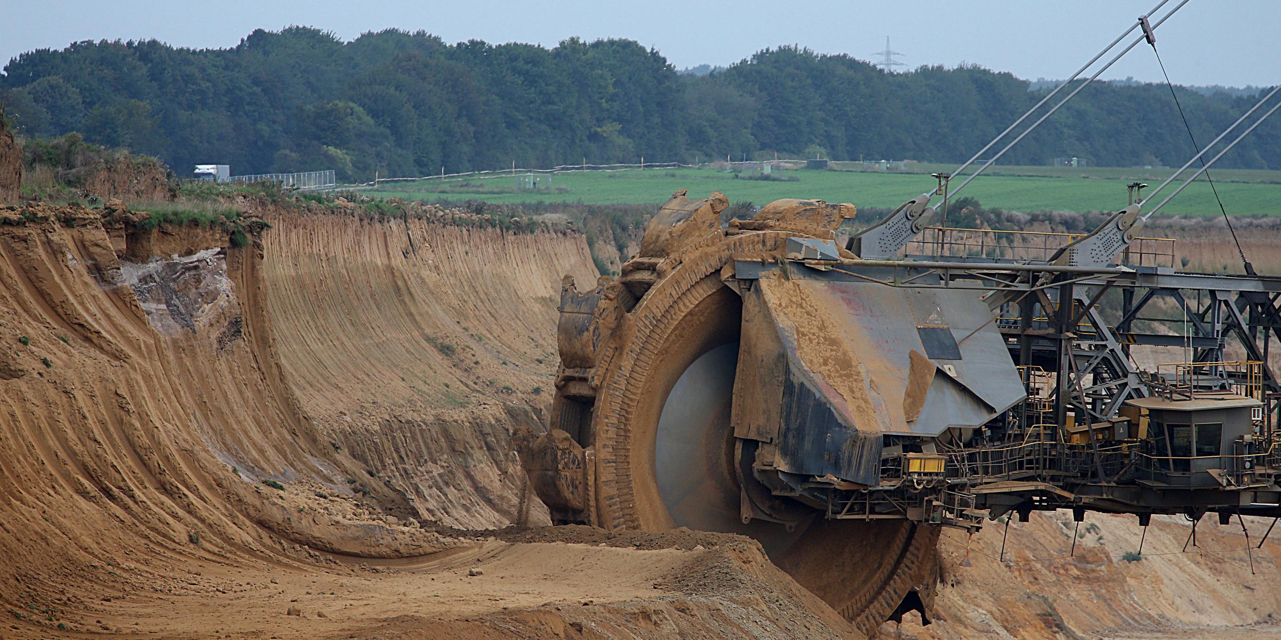 Ein Schaufelrad-Bagger gräbt Erde weg. Im Hintergrund ein Wald und Grasland