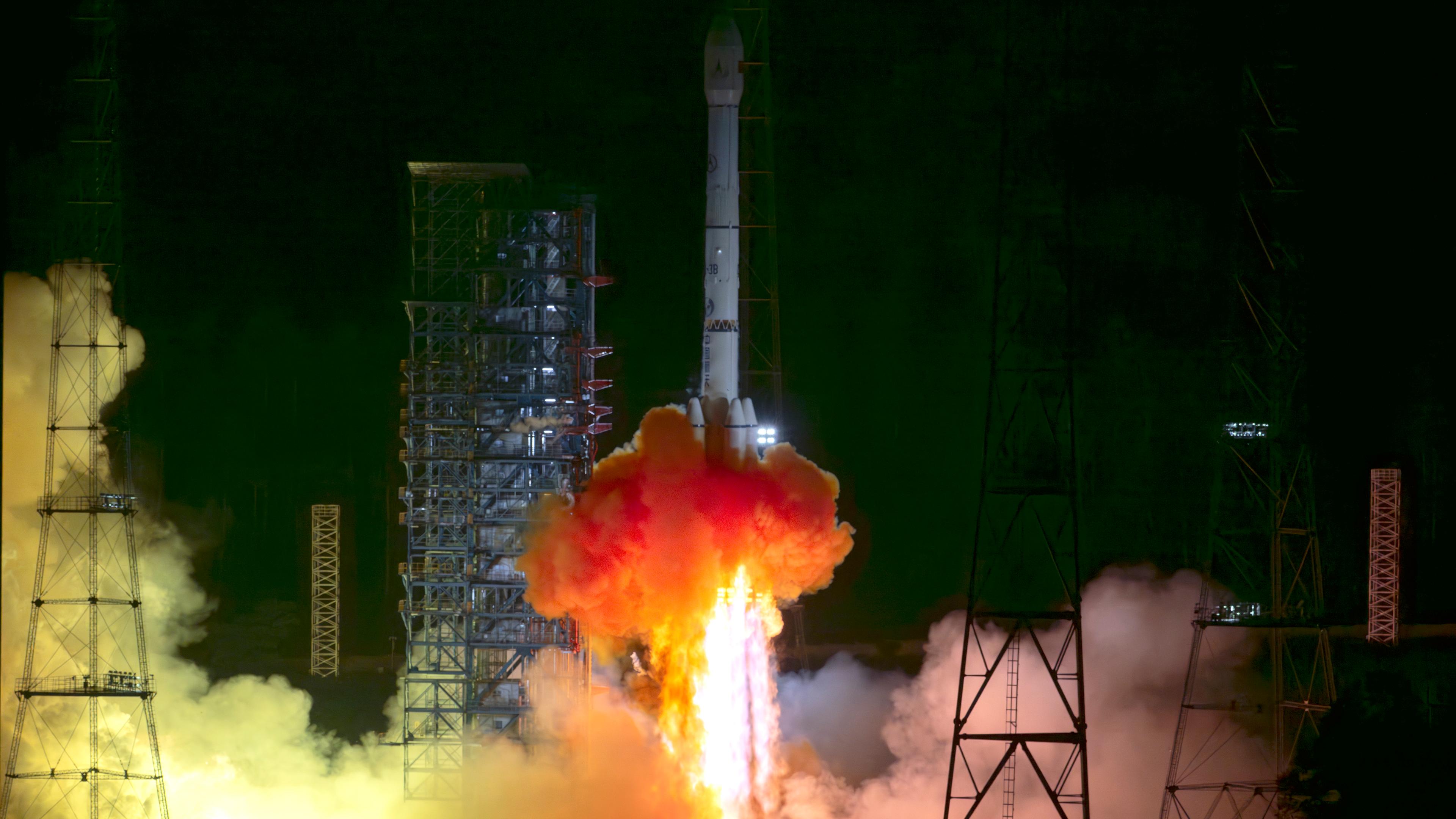 Eine Rakete startet bei Nacht. Aus der unteren Öffnung schießen helle Flammen und Rauchwolken in Gelb, Orange und Weiß. Die Umgebung ist dunkel, der Startplatz von Scheinwerfern beleuchtet.