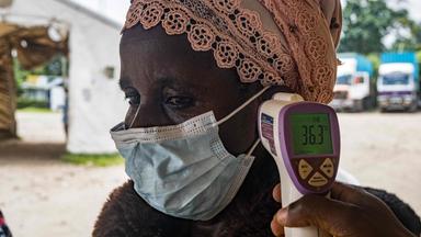 Frau mit Mundschutz bekommt ein Fieberthermometer an die Stirn. 