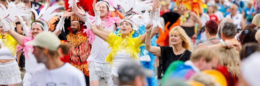 Viele Menschen ziehen gemeinsam durch die Stadt Köln. Einige tragen bunte Kostüme mit großen Federn, andere schwenken Regenbogenflaggen und feiern fröhlich zusammen.
