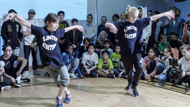  Im Vordergrund tanzen Dascha und Rocco, hinter ihnen sieht man einen Teil des Publikums – ca. 20 Kinder und Jugendliche, die zum Teil mitklatschen. Dascha und Rocco tragen dunkelblaue T-Shirts mit der Aufschrift „Limited Edition“ und führen gerade einen synchron Breakdance-Move aus, dem sie die Arme heben, und die Füße überkreuzen.