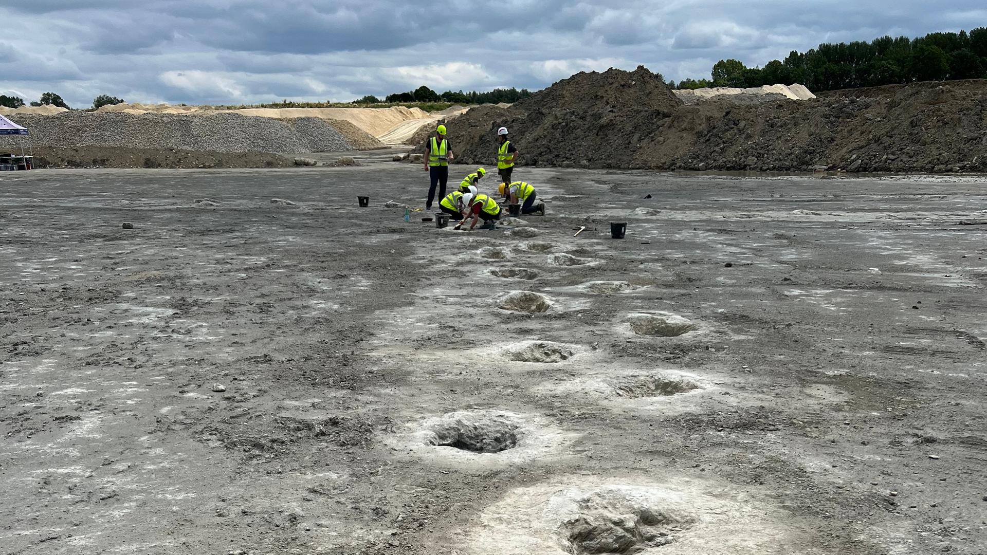 Regelmäßige Löcher im Boden, im Hintergrund Menschen in Warnwesten, die über einem Loch knieen.