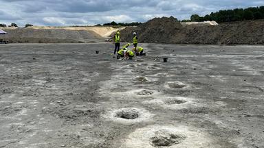 Regelmäßige Löcher im Boden, im Hintergrund Menschen in Warnwesten, die über einem Loch knieen.