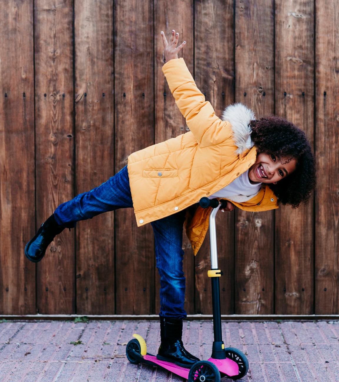 Ein Mädchen mit braunen Locken steht grinsend auf einem Skateboard und streckt ein Bein und einen Arm raus. Sie trägt eine gelbe Jacke und schwarze Gummistiefel.