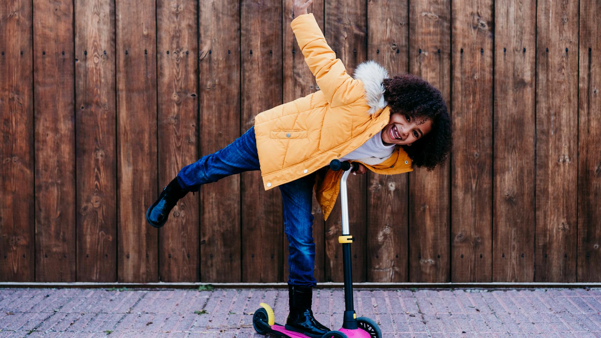 Ein Mädchen mit braunen Locken steht grinsend auf einem Skateboard und streckt ein Bein und einen Arm raus. Sie trägt eine gelbe Jacke und schwarze Gummistiefel.