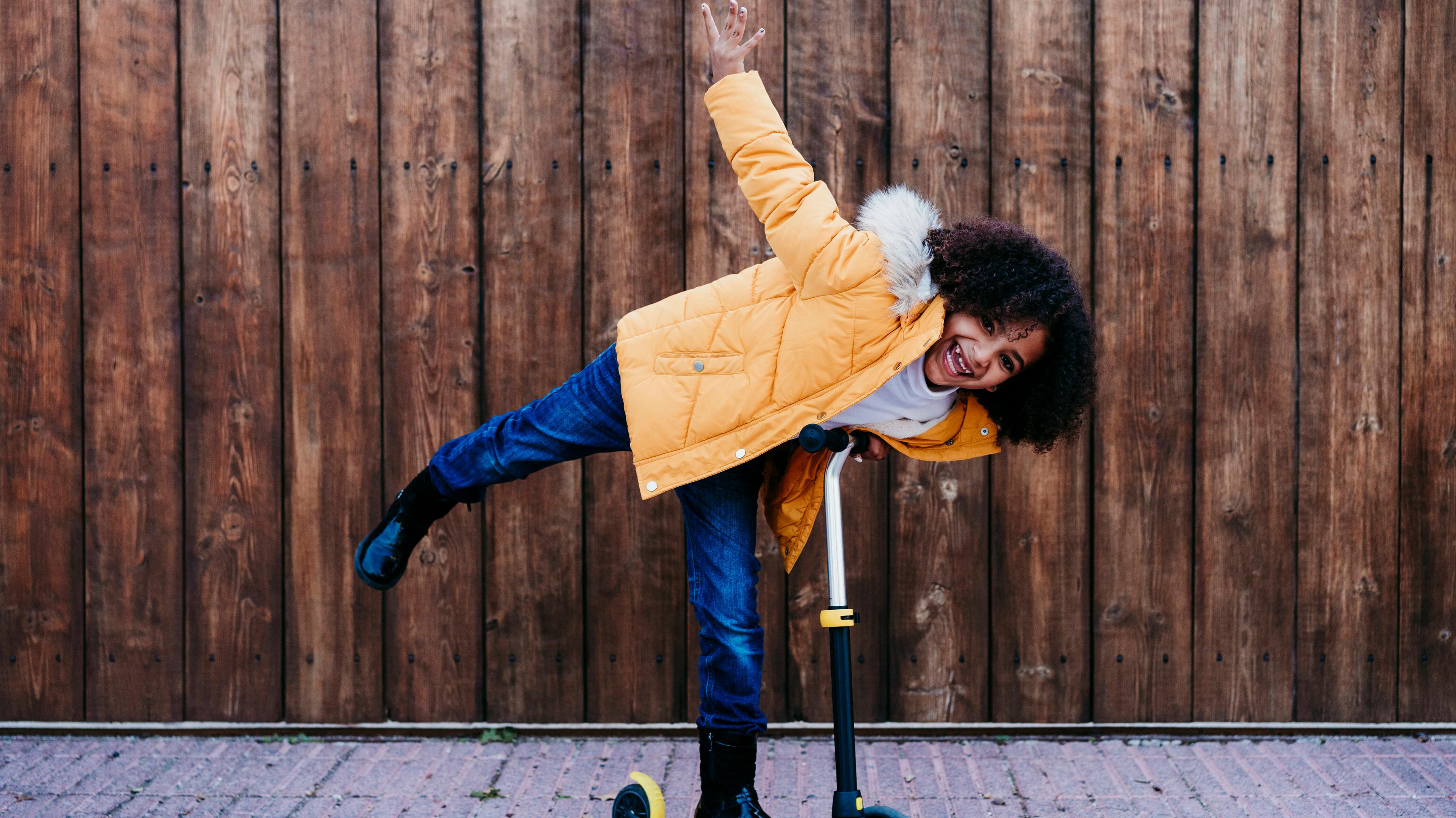 Ein Mädchen mit braunen Locken steht grinsend auf einem Skateboard und streckt ein Bein und einen Arm raus. Sie trägt eine gelbe Jacke und schwarze Gummistiefel.
