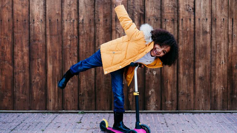 Ein Mädchen mit braunen Locken steht grinsend auf einem Skateboard und streckt ein Bein und einen Arm raus. Sie trägt eine gelbe Jacke und schwarze Gummistiefel.