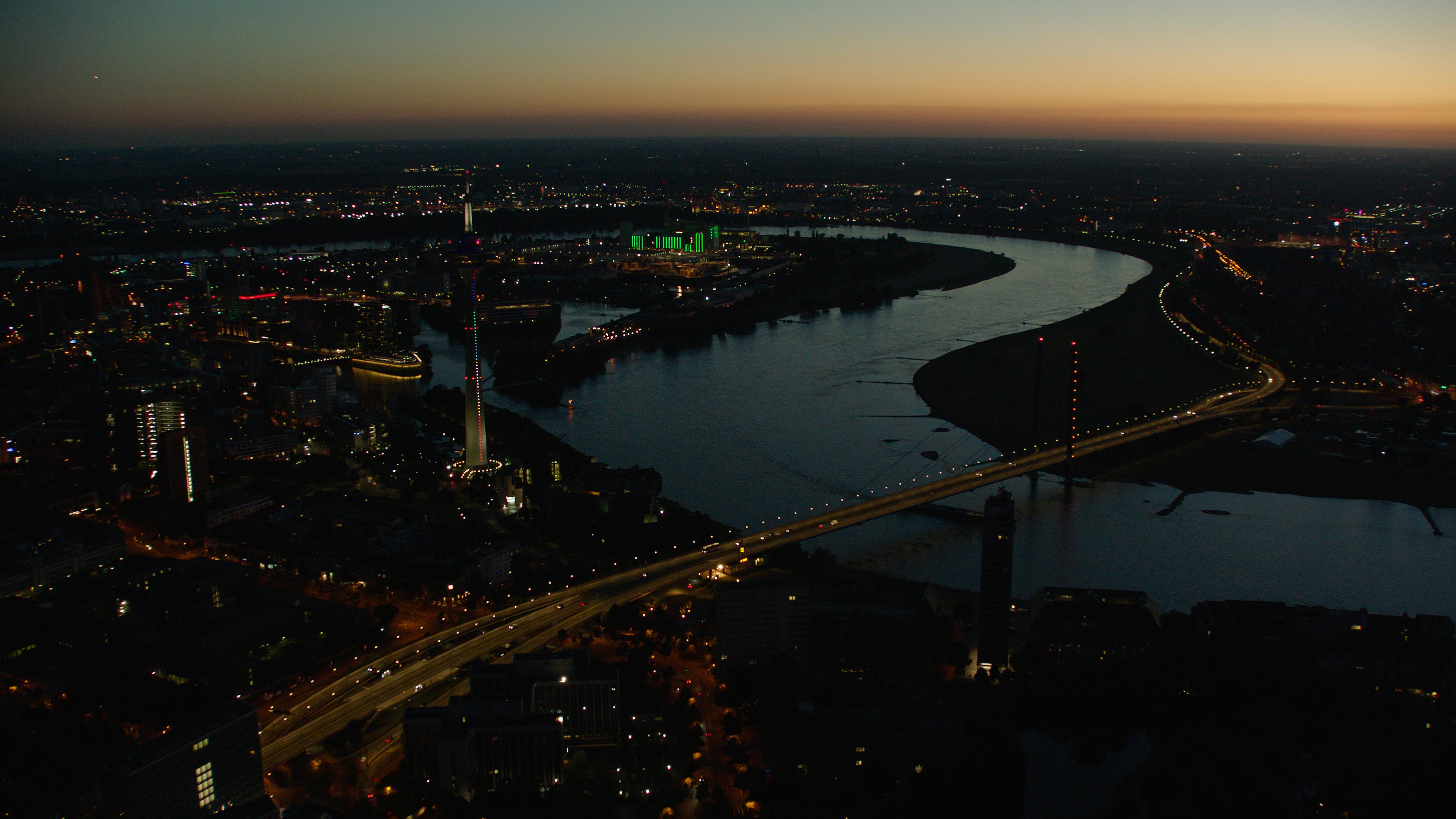 Luftaufnahme von Düsseldorf. Deutlich erkennbar der Rhein. Und vorne im Bild eine beleuchtete Brücke über den Rhein.