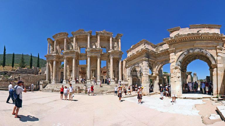 Ruinen einer antiken Bibliothek in Ephesus in der Türkei. Strahlendblauer Himmel, Touristen schauen sich die Säulen, Treppen und Fassadenteile an.