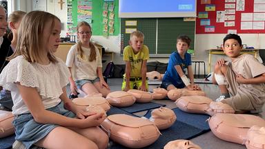 Kinder sitzen im Klassenzimmer um Puppen herum, bei einem Erste Hilfe Kurs.
