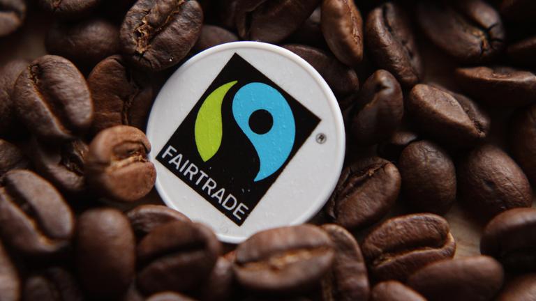 Auf dem Foto sieht man einen Haufen an dunkelbraunen Kaffeebohnen, in denen eine Münzgroße Plakette liegt. Auf dieser kleinen Münze, bzw. weißen Plakette, ist das blau, grün und schwarze Fairtrade-Logo draufgedruckt.