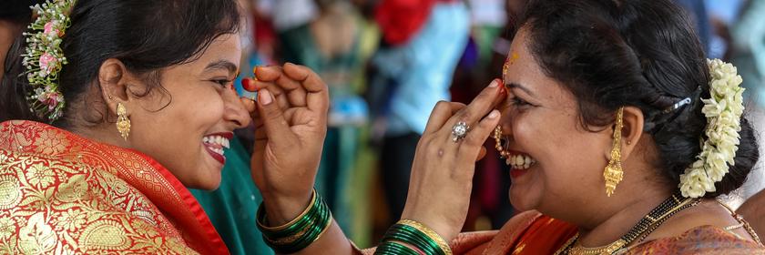 Zwei Frauen in traditioneller indischer Kleidung malen sich gegenseitig einen Punkt auf die Stirn.