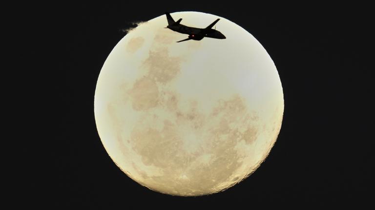 Ein Flugzeug fliegt am Himmel. Im Hintergrund ist der Mond zu sehen. 