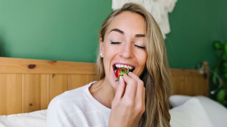 Eine Frau verspeist genüsslich eine Erdbeere in ihrem Bett. 