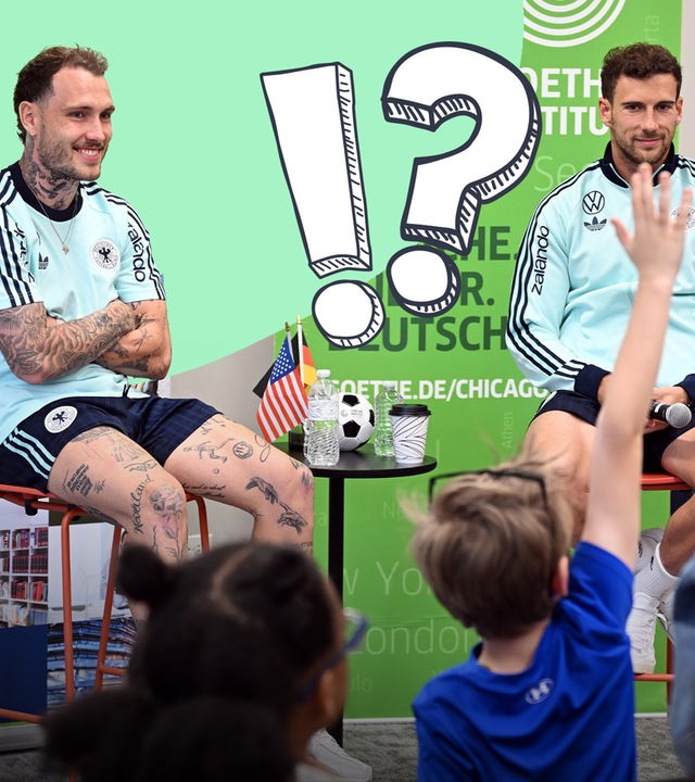 Goretzka und Raum sitzen auf Barhockern vor Kindern, die die Hände heben. Zwischen ihnen zwei grafische Symbole, ein Fragezeichen und ein Ausrufezeichen.