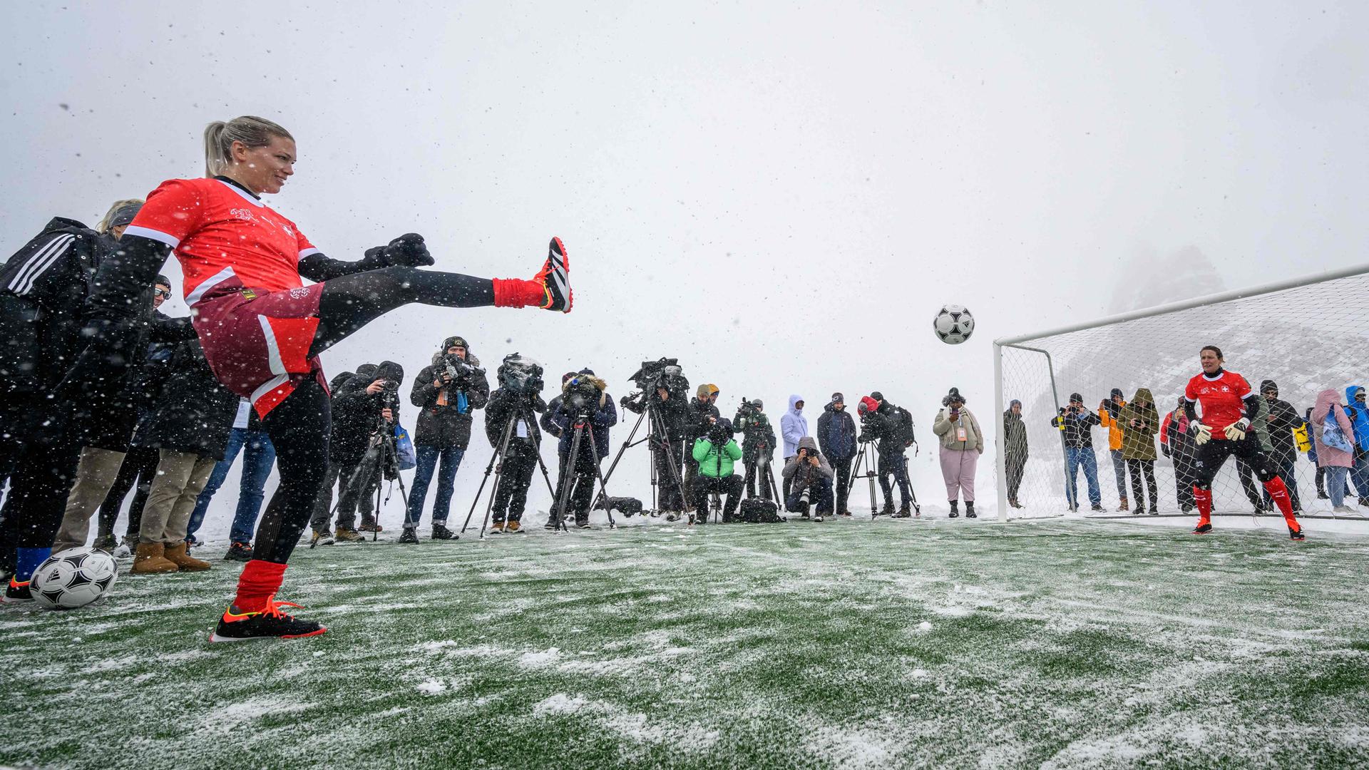 Lara Dickenmann schießt auf ein Tor, im Hintergrund ist ein verschneiter Berg zu sehen. Rund um das Fußballfeld stehen Kameraleute.
