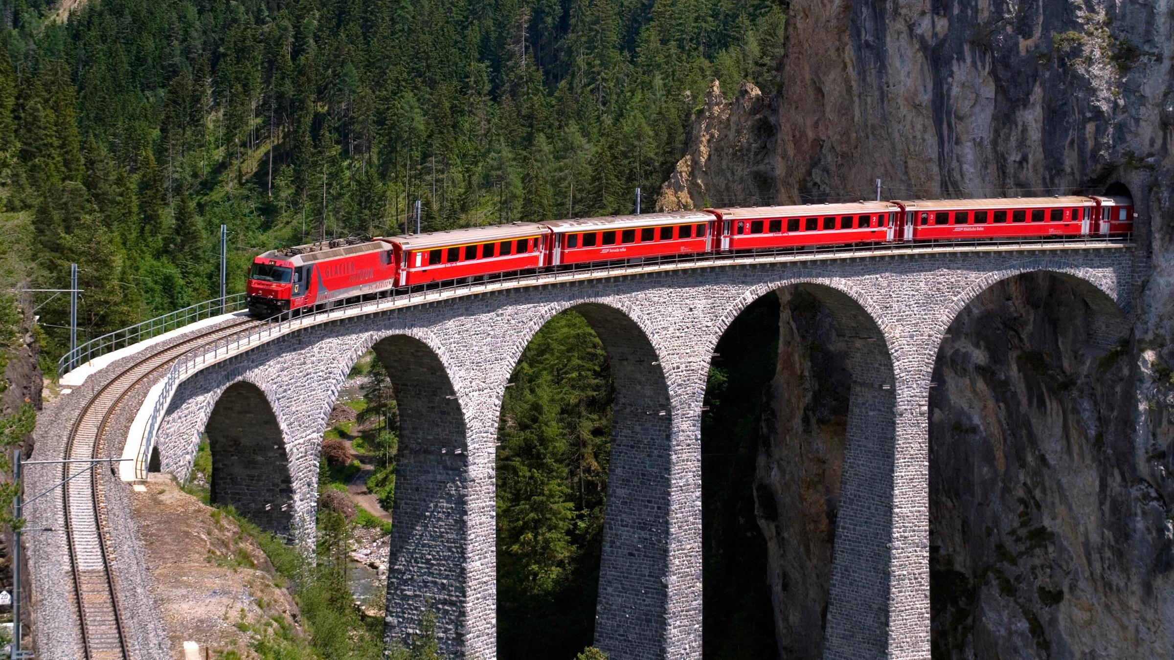 Ein roter Zug fährt aus einem Tunnel direkt auf eine hohe Brücke.