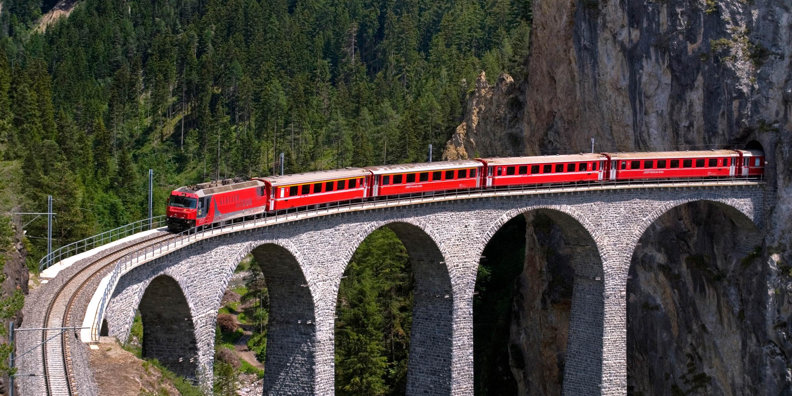 Ein roter Zug fährt aus einem Tunnel direkt auf eine hohe Brücke.