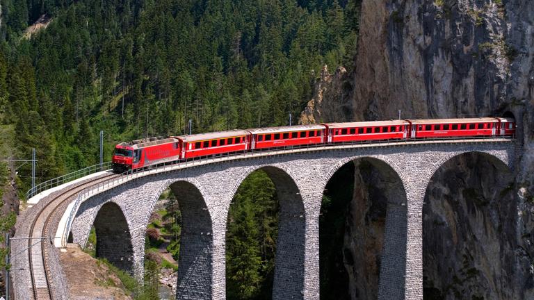 Ein roter Zug fährt aus einem Tunnel direkt auf eine hohe Brücke.