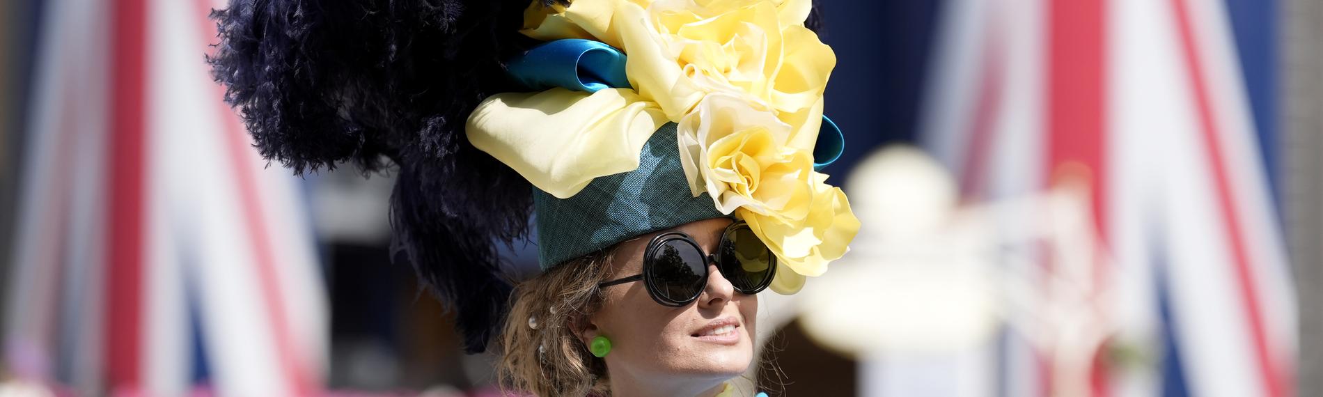 Eine Besucherin des "Royal Ascot" in Großbritannien mit einem riesigen, bunten Hut aus Federn und Stoffblumen vor zwei britischen Flaggen.
