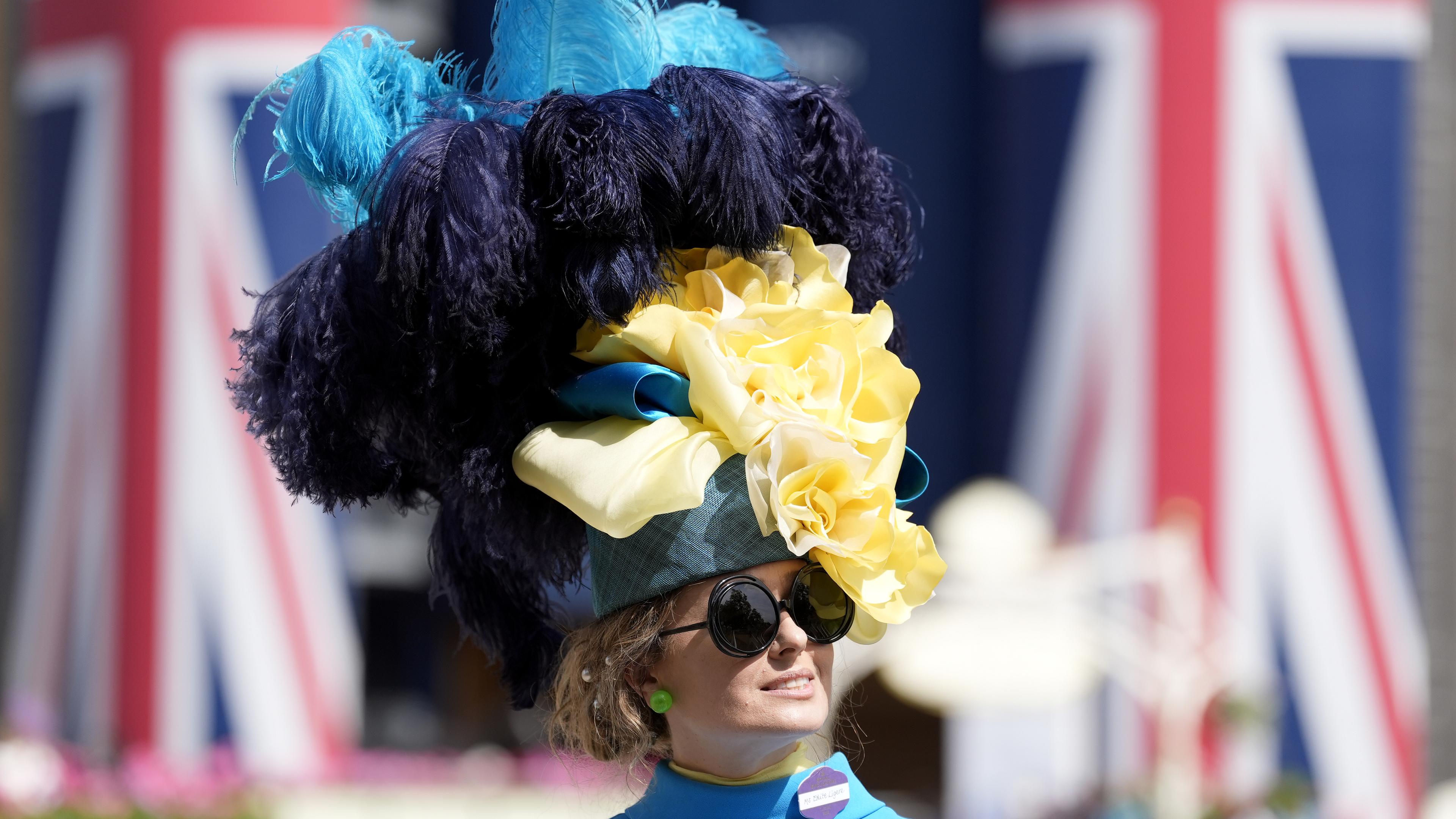 Eine Besucherin des "Royal Ascot" in Großbritannien mit einem riesigen, bunten Hut aus Federn und Stoffblumen vor zwei britischen Flaggen.