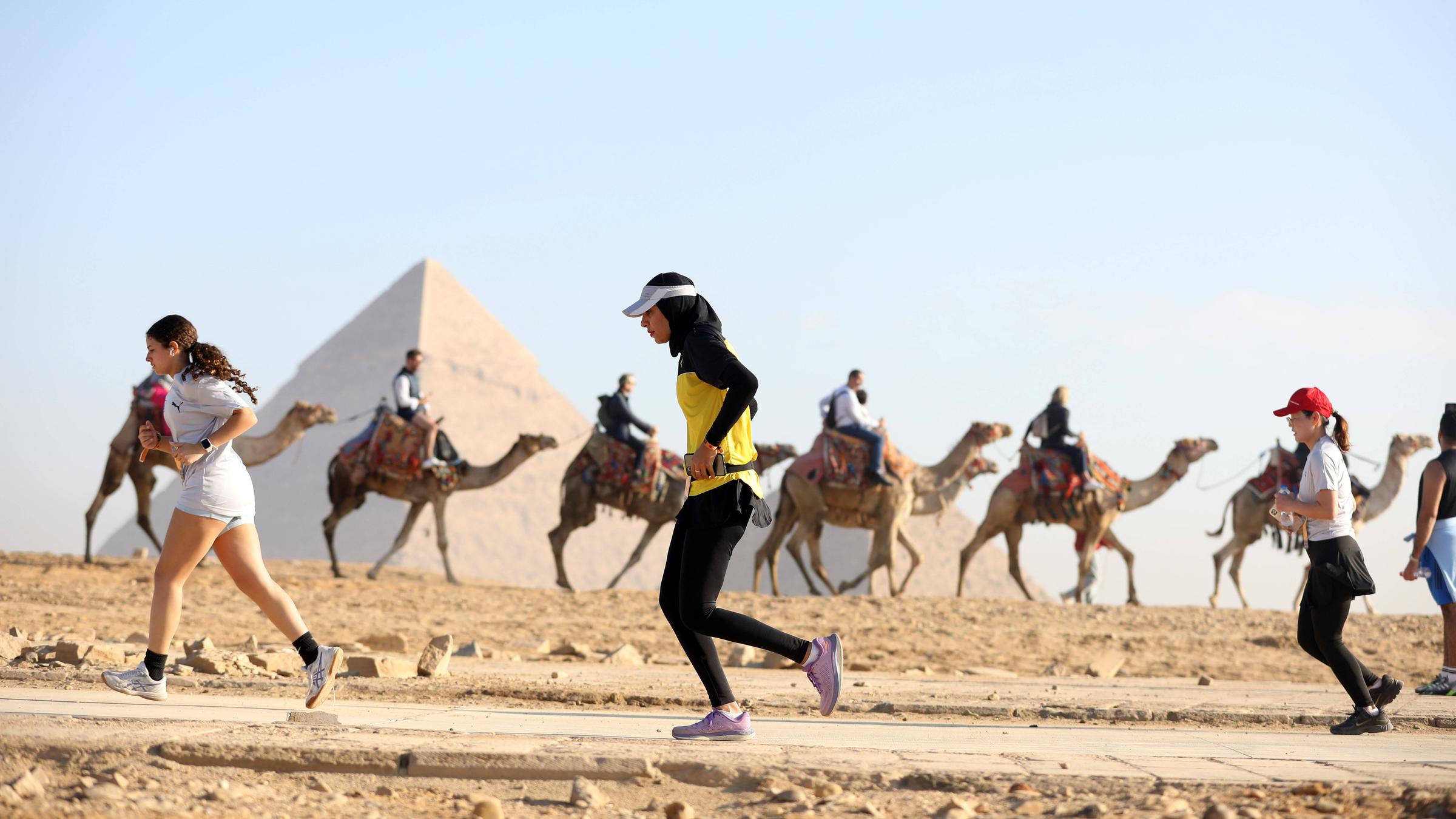 Im Vordergrund laufen Sportlerinnen und Sportler über eine Straße durch die Wüste. Im Hintergrund ragen die Pyramiden auf, während Kamele mit Reitern daneben entlangziehen.