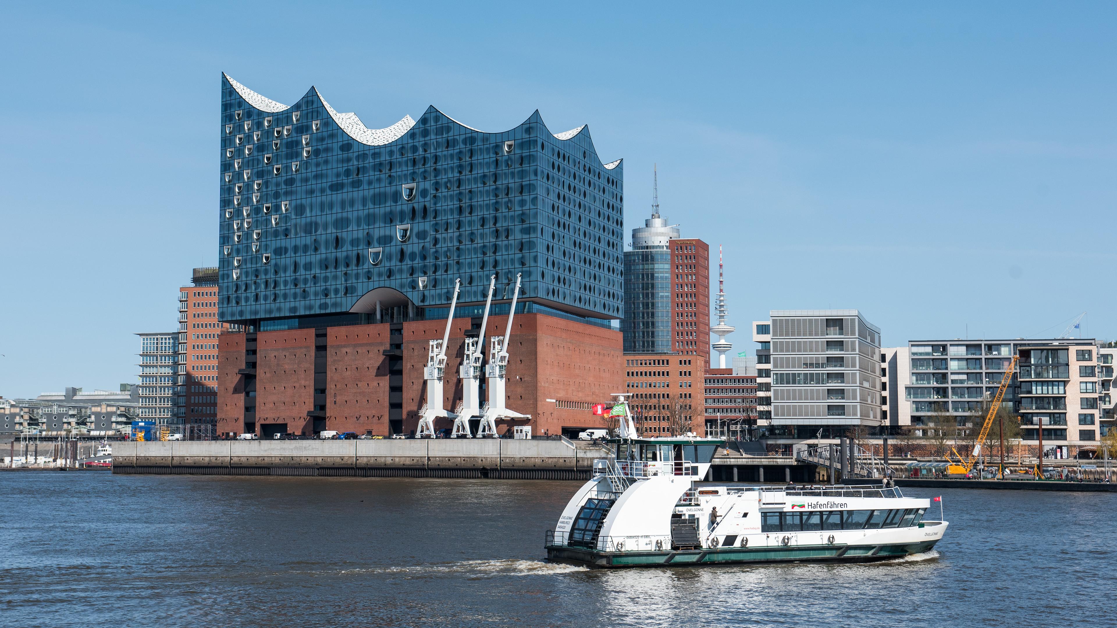Eine Hafenfähre ist bei blauem Himmel und Sonnenschein auf der Elbe vor der Elbphilharmonie unterwegs. 