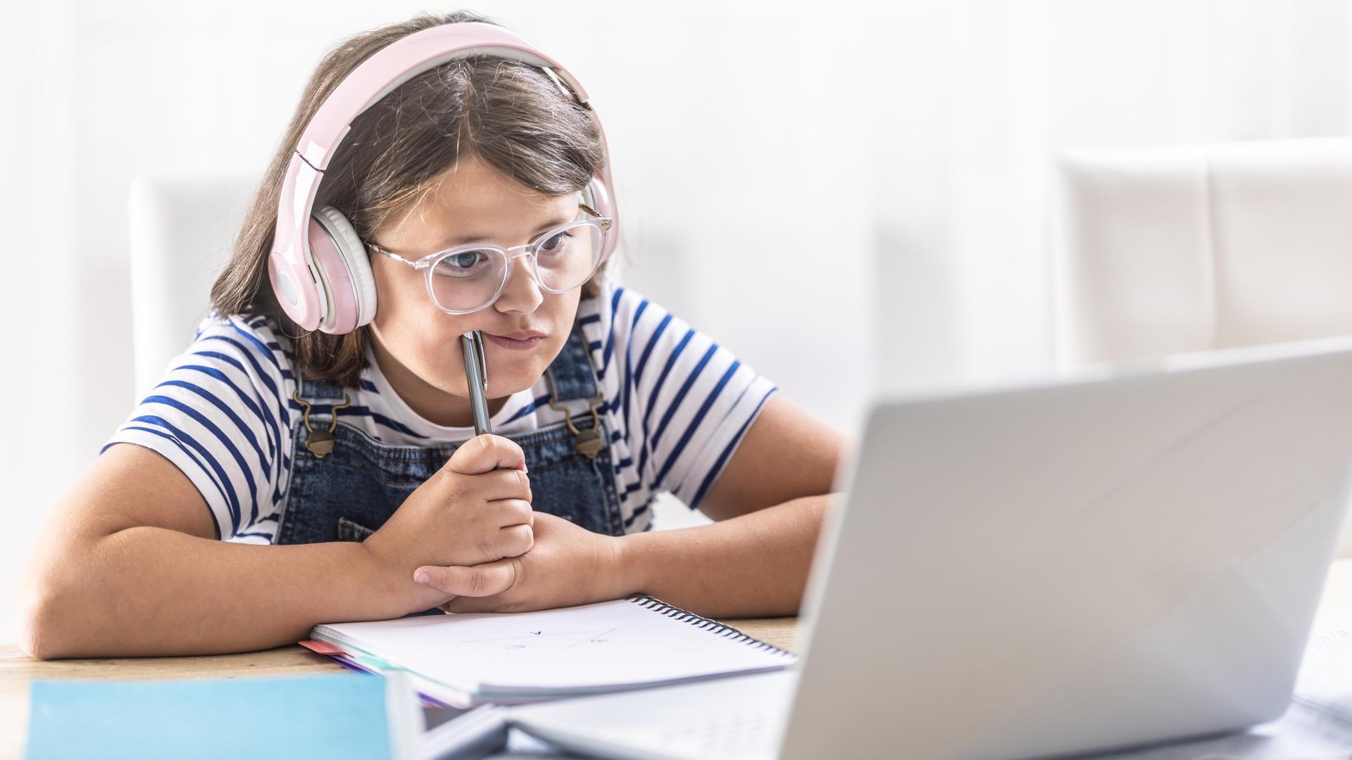 Mädchen mit Kopfhörern grübelt über Papier vor einem Laptop und erledigt ihre Hausaufgaben.